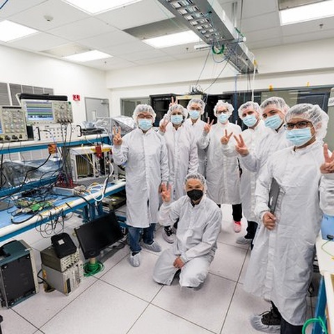 An electronics laboratory with oscilloscopes and instrument racks along one wall, and a group of engineers in laboratory coats.