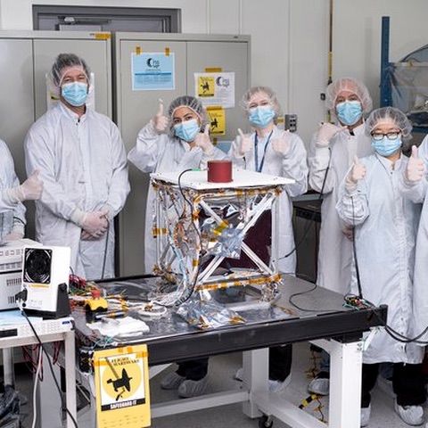 An engineering group in laboratory coats and hairnets gathered around a foil-wrapped truss assembly on an anti-static bench.
