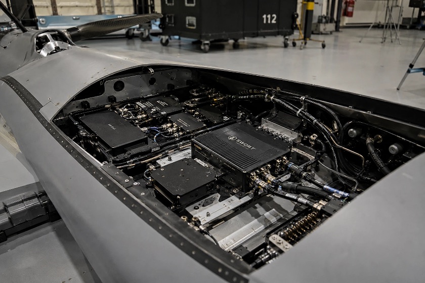 An open avionics bay in an airframe, showing line-replaceable electronics modules, coaxial and harness runs, and connector backshells on a mounting tray.